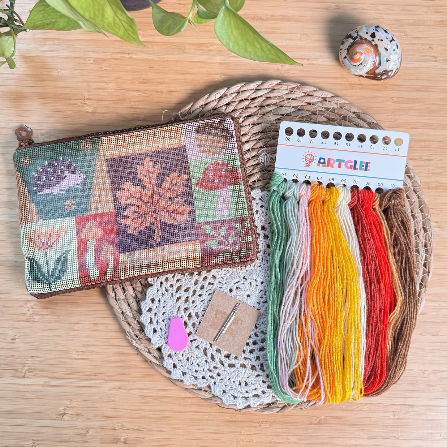 Autumn Forest Half-Stitch Bag Kit flat lay, featuring a patchwork embroidered pouch with autumn leaves, mushrooms, and woodland motifs, shown with embroidery threads, needle, and tools on a wooden surface.