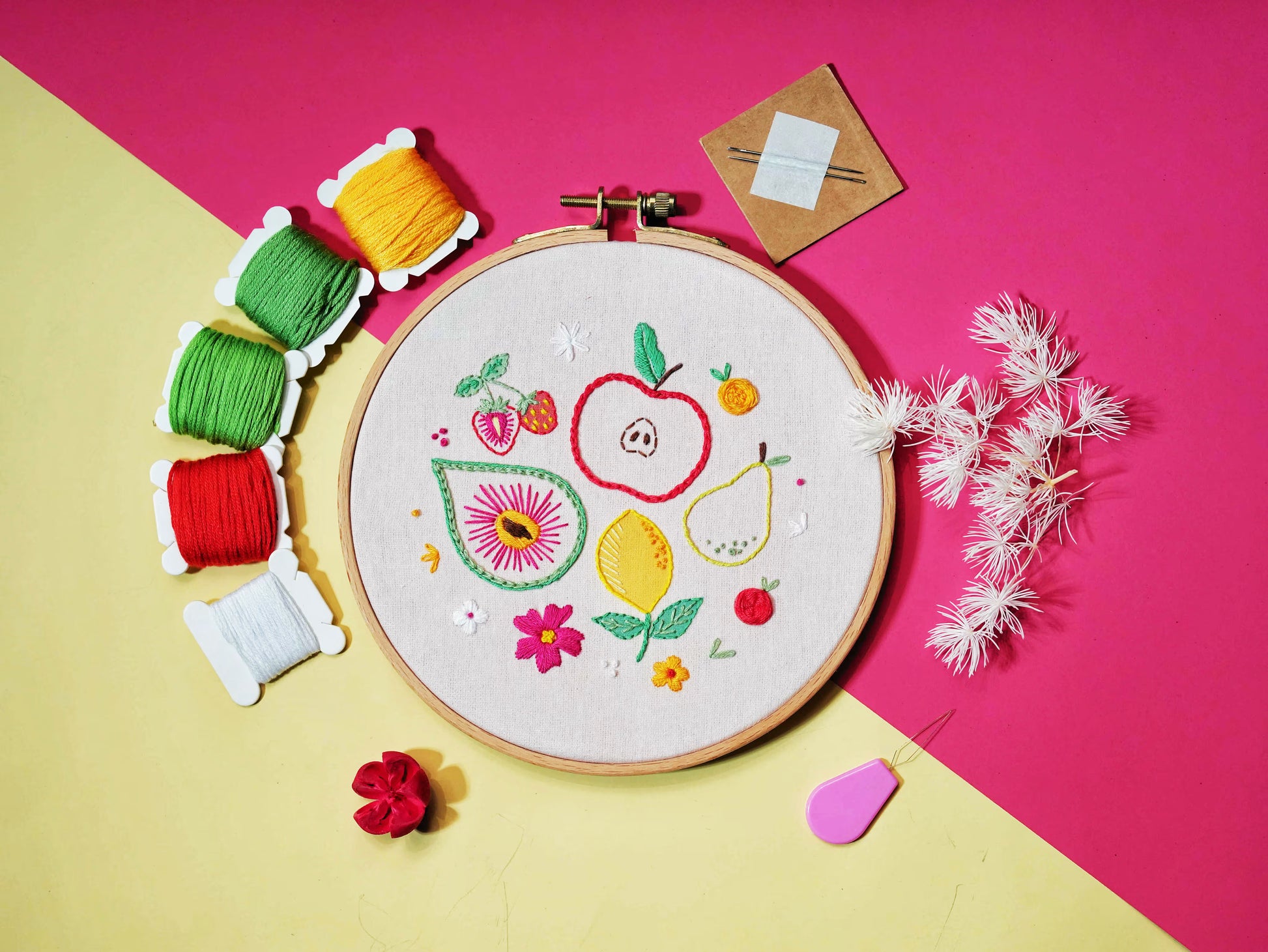 Flat lay of Fruity Fiesta Embroidery Kit, including embroidery hoop with fruit and flower patterns, colorful threads, needles, and accessories on a bright pink and yellow background.
