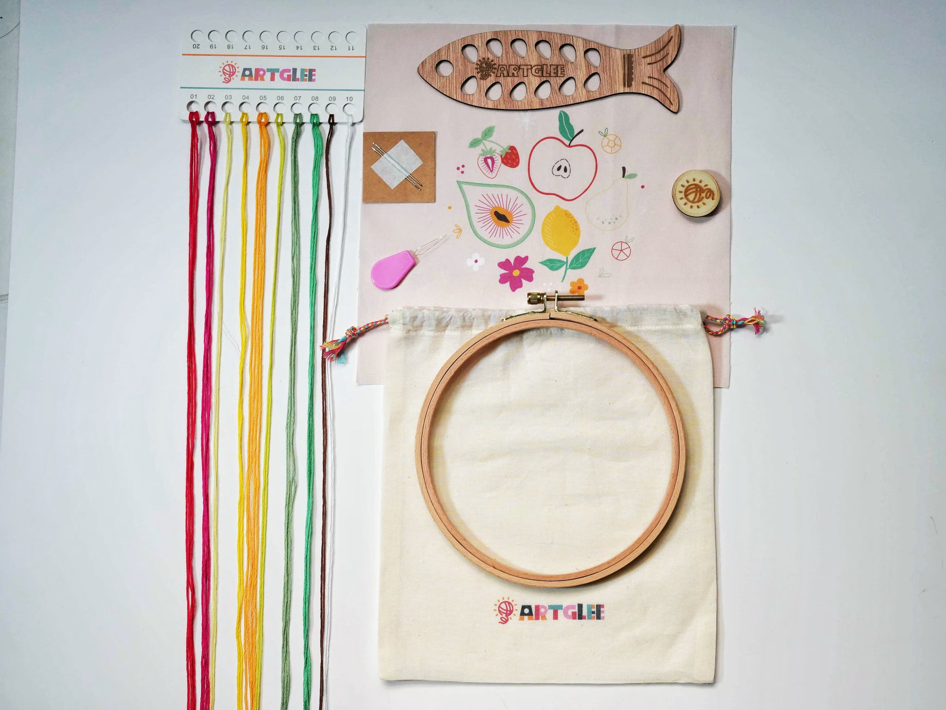 Complete Fruity Fiesta Embroidery Kit set with printed fabric, colorful threads on fish-shaped thread holder, wooden embroidery hoop, needles, threader, and drawstring storage bag.