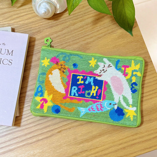 Completed Lucky Cats half-stitch zipper pouch, laid flat on a wooden table with green fabric background and playful “I’m Rich” cat design.