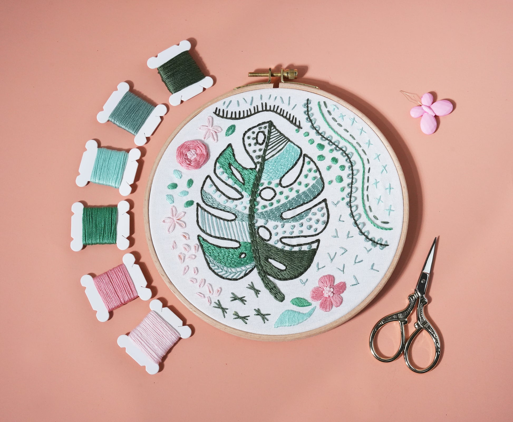 Monstera embroidery beginner kit with green leaf hoop, colorful threads, and scissors on pink background.
