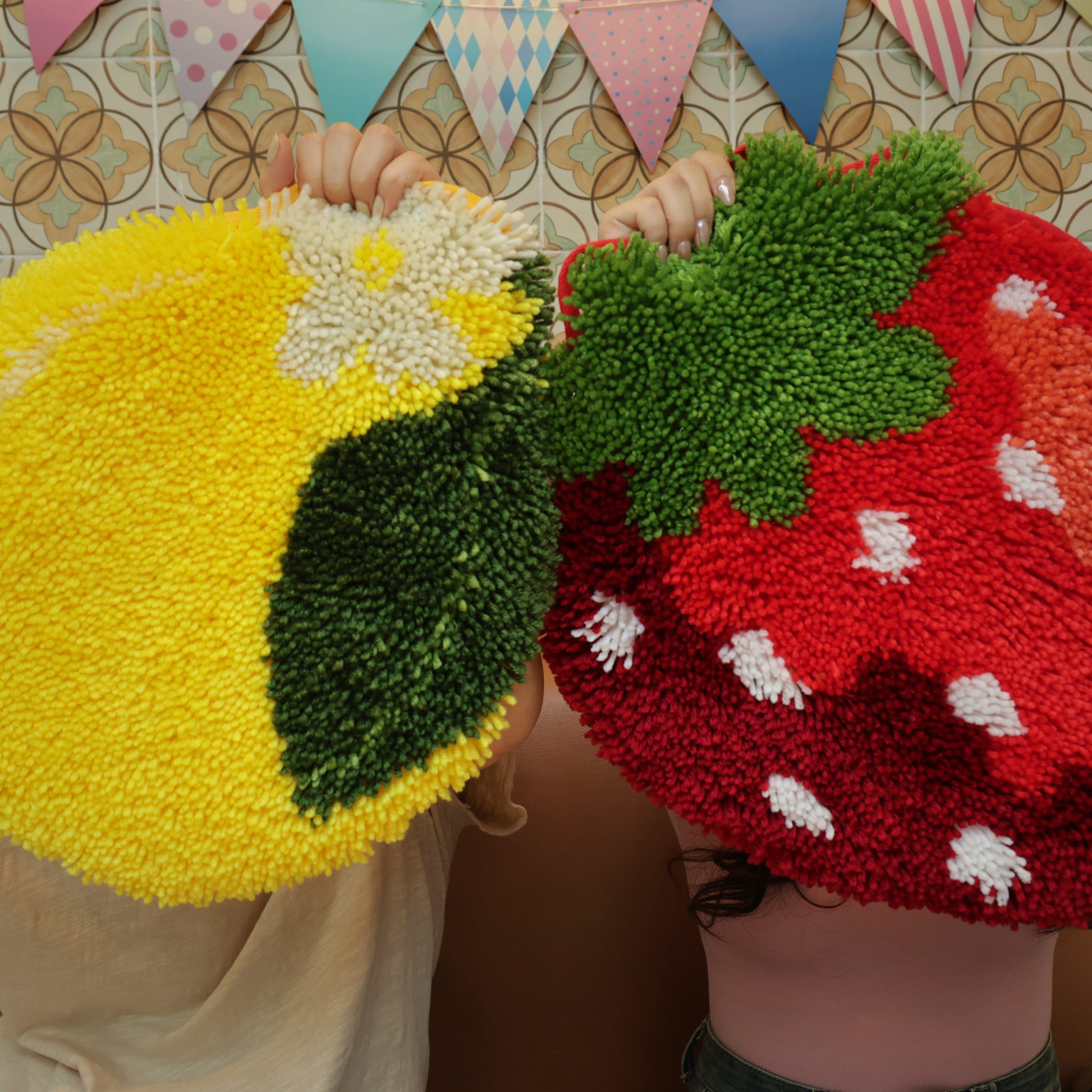 Two people playfully holding up handmade latch hook rugs shaped like a bright yellow lemon and a vibrant red strawberry, showcasing the Sweet Strawberry Latch Hook Kit in a fun and colorful display.