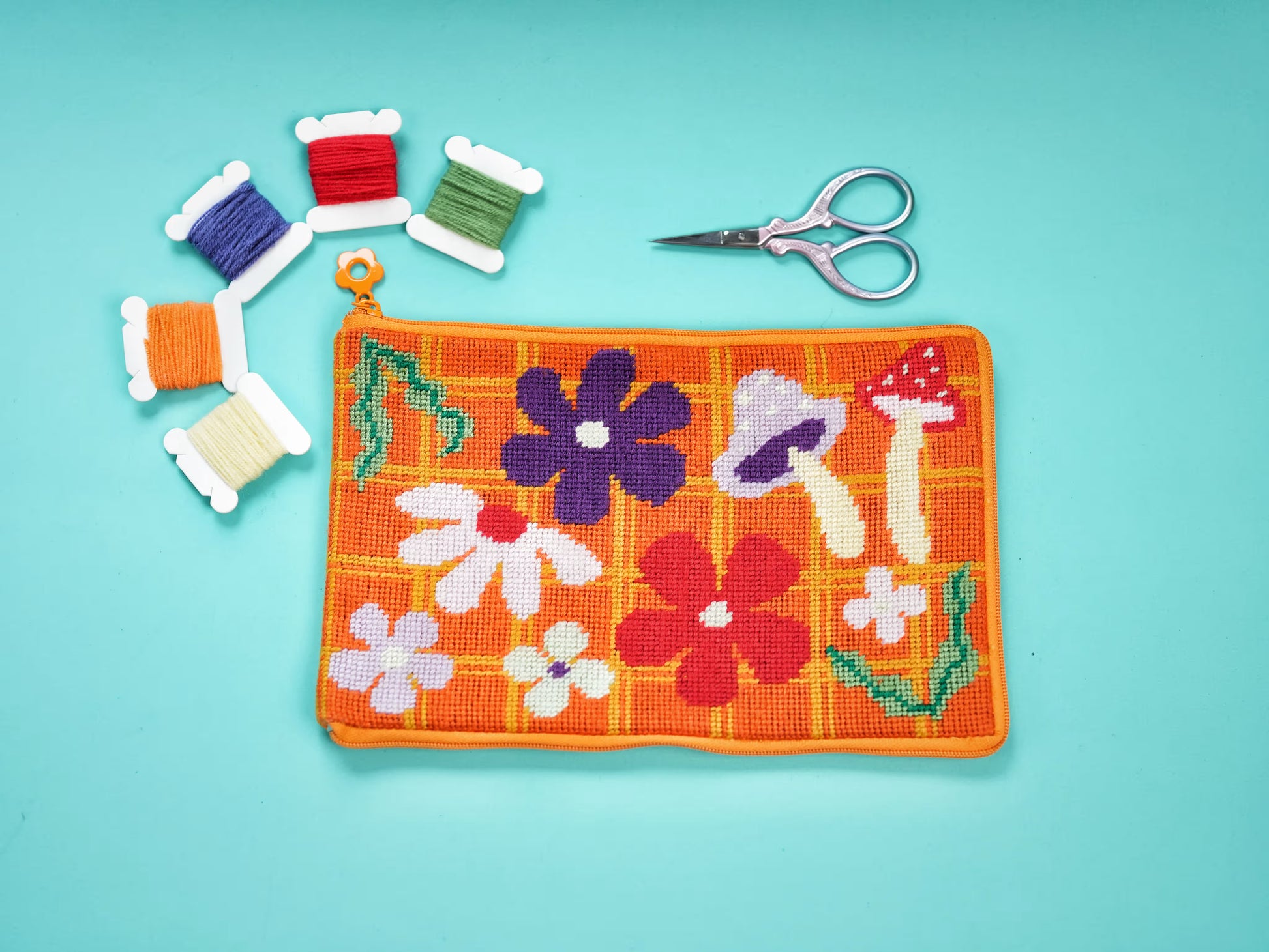 Bloom Daisy Half-Stitch Kit pouch laid flat on a turquoise background, featuring colorful floral and mushroom motifs, embroidery threads, and scissors.
