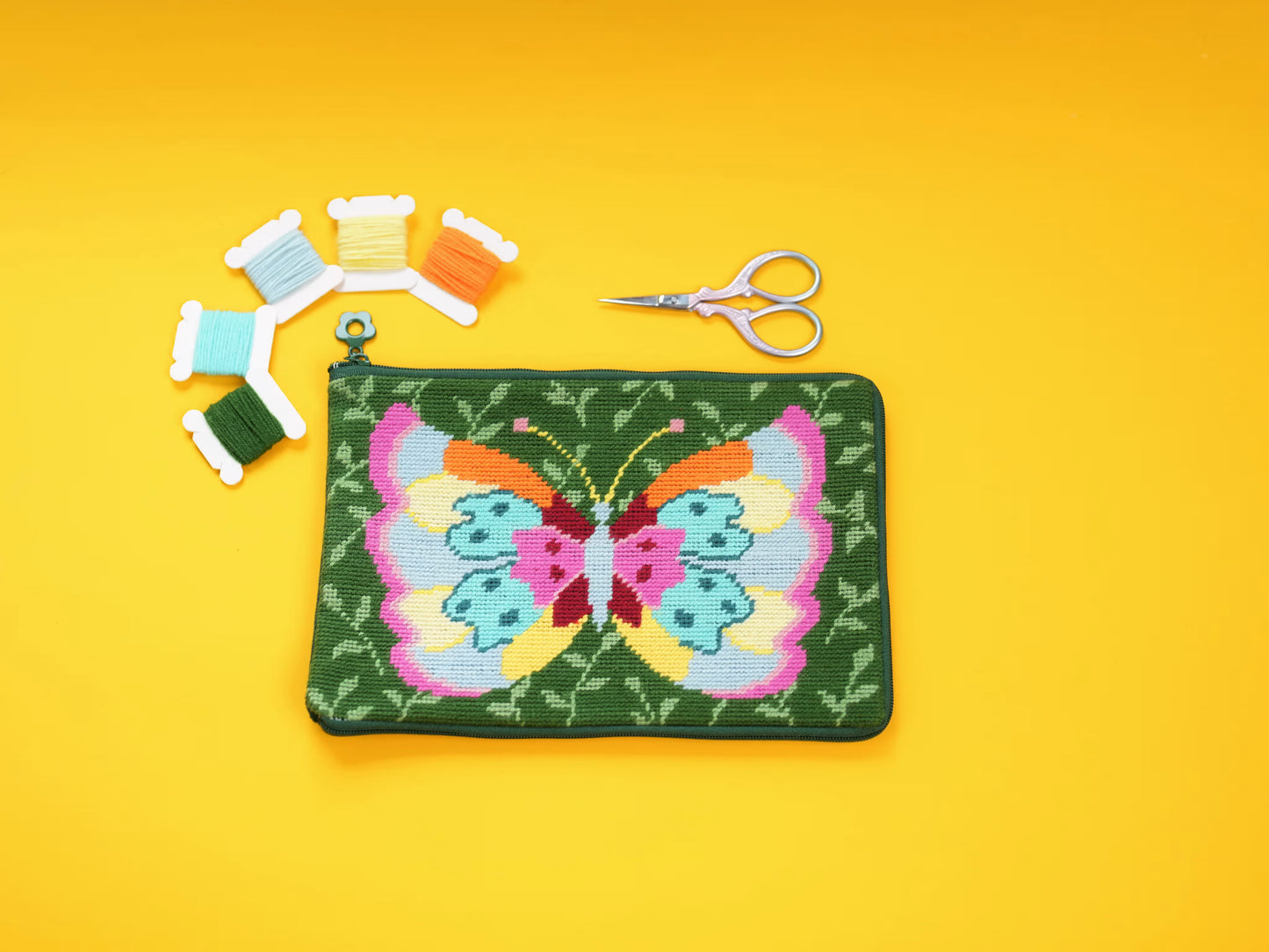 Butterfly Garden Half-Stitch Kit pouch laid flat on a yellow background, featuring a colorful butterfly design with embroidery threads and scissors.
