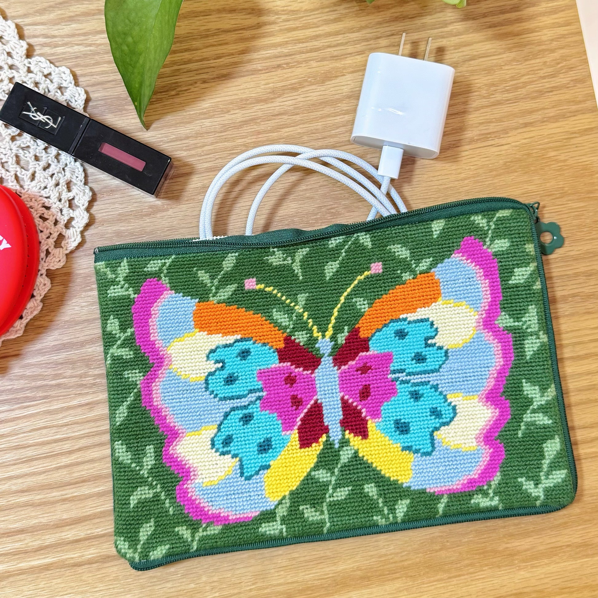 Butterfly Garden Half-Stitch Kit pouch used as storage for makeup and charger, placed on a wooden table.