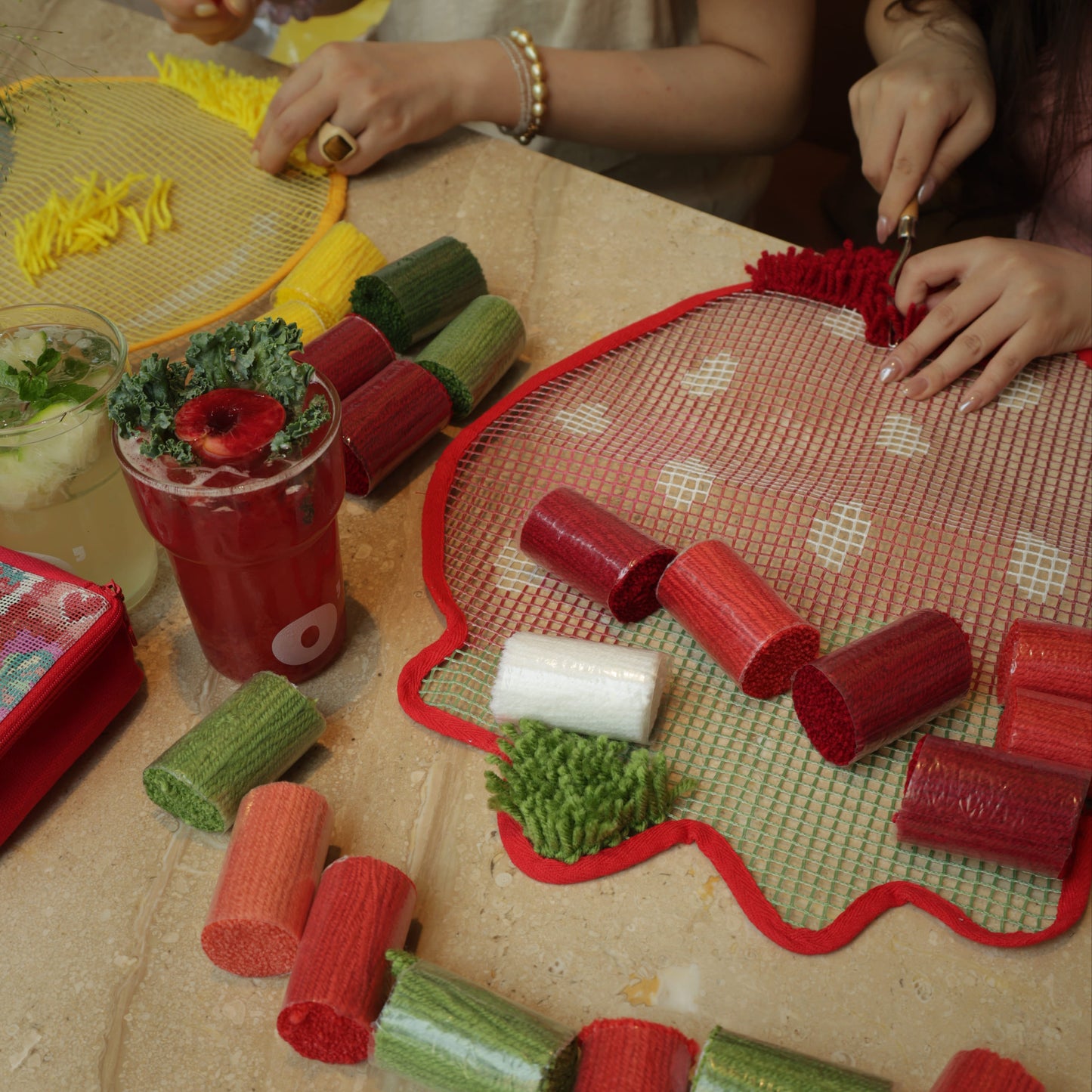 DIY crafting scene with two people working together on strawberry and lemon latch hook kits, colorful yarn and drinks on the table.