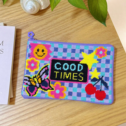 Flatlay of the Good Times Half-Stitch Kit finished pouch with retro smiley, butterfly, flowers, stars, and cherries on a purple checkered background.
