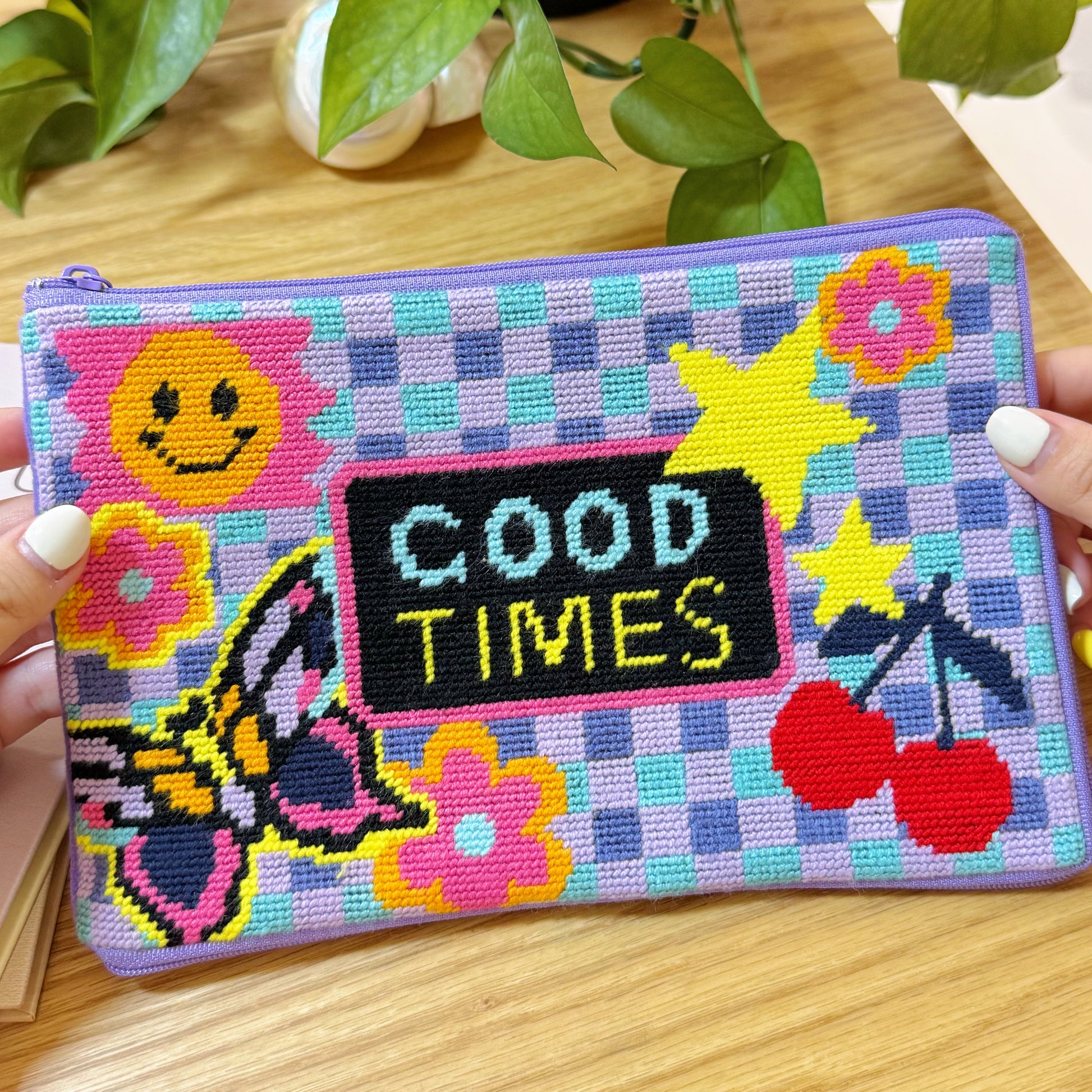 Good Times Half-Stitch Kit pouch held in hands, showing colorful stitched motifs with smiley face, stars, cherries, and butterfly.