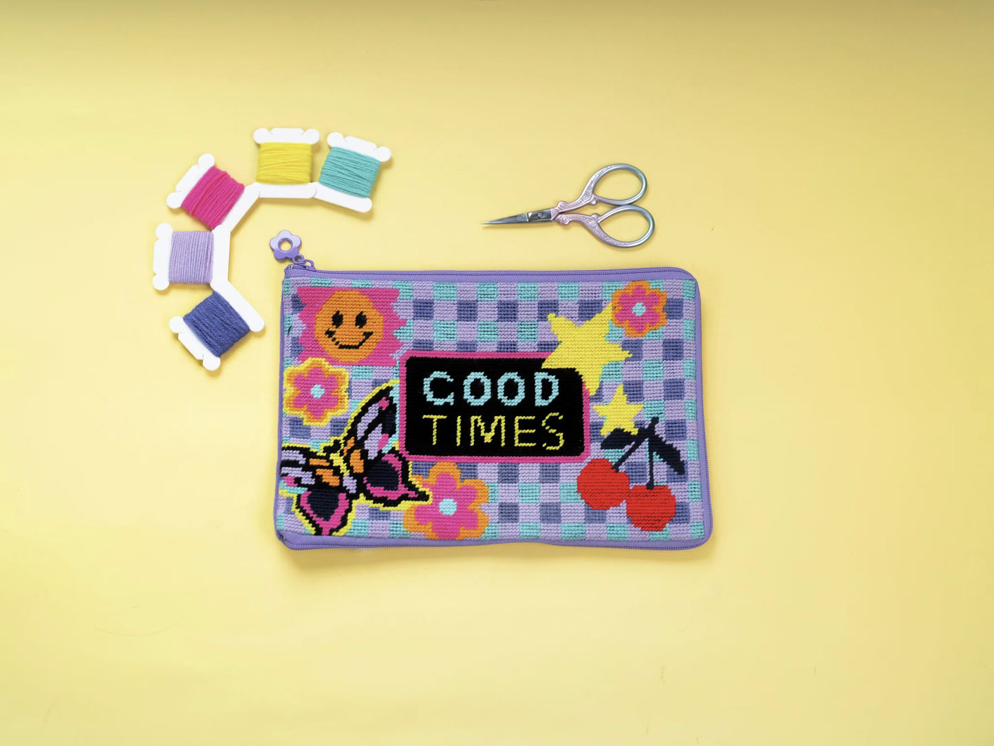 Good Times Half-Stitch Kit pouch laid flat on a yellow background, featuring a playful checkerboard design with text, butterflies, flowers, embroidery threads, and scissors.

