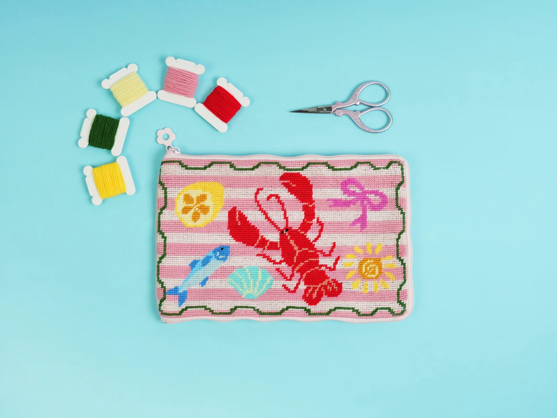 Lucky Catch Coast Half-Stitch Kit flat lay, featuring an embroidered pouch with a red lobster, fish, seashells, and coastal motifs on a soft blue background.
