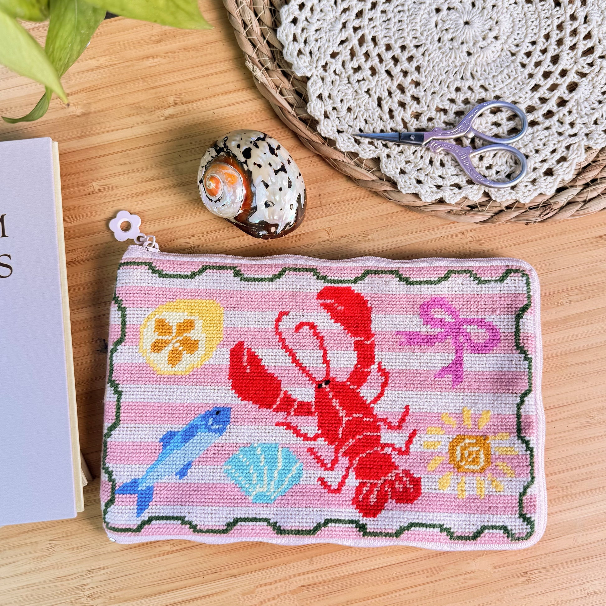 Lucky Catch Coast Half-Stitch Kit flat lay, featuring a coastal lobster embroidered pouch with fish and shell motifs, styled on a wooden surface with scissors, shell decor, and craft accessories.
