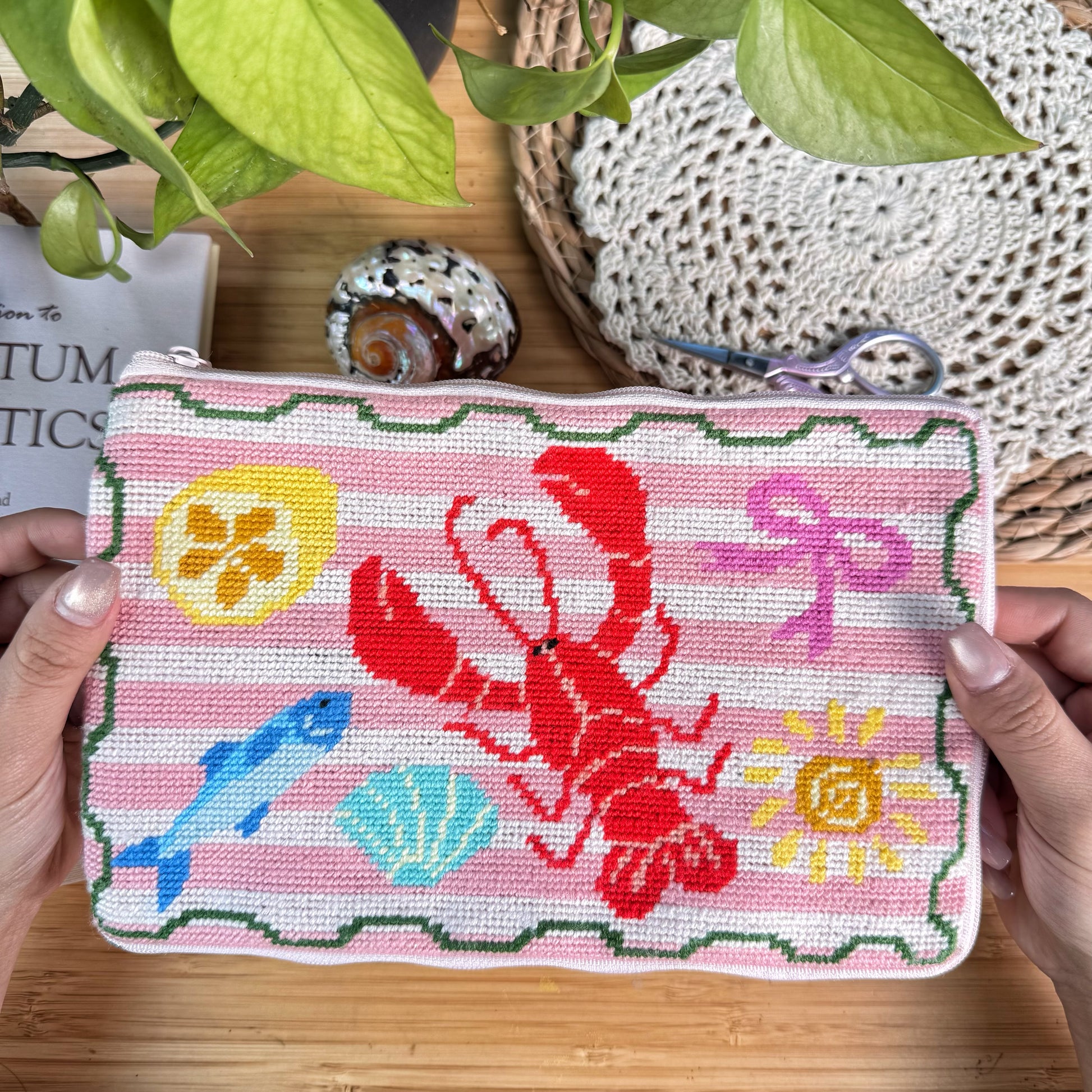 Lucky Catch Coast Half-Stitch Kit held in hands, featuring a coastal lobster embroidered pouch with fish and shell motifs, styled on a wooden surface with natural decor.
