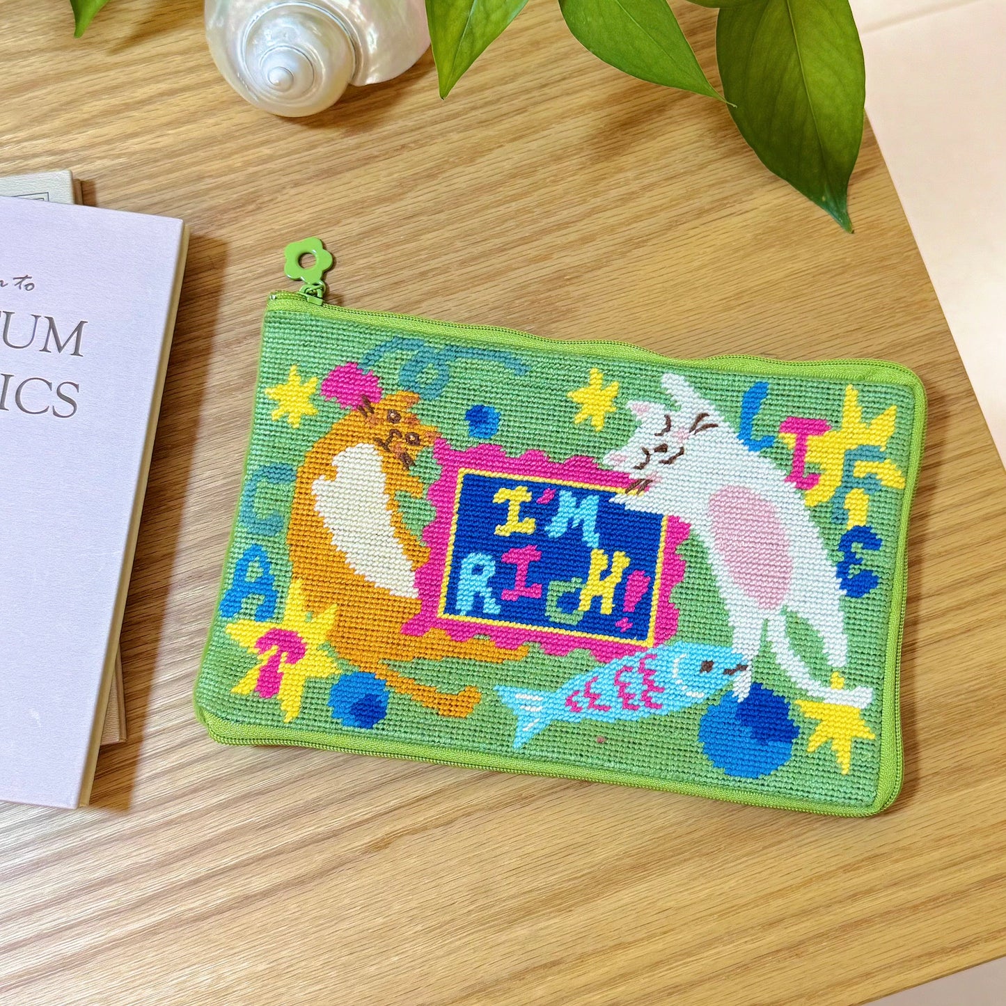 Completed Lucky Cats half-stitch zipper pouch, laid flat on a wooden table with green fabric background and playful “I’m Rich” cat design.