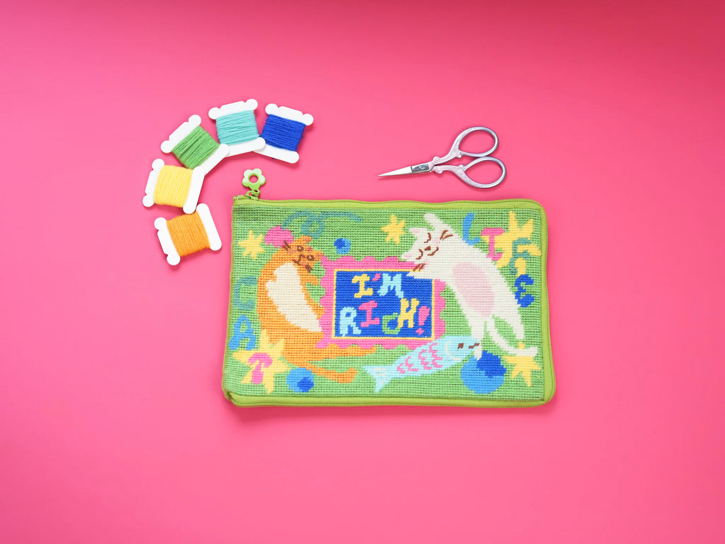 Lucky Cats Half-Stitch Kit pouch laid flat on a pink background, featuring colorful lucky cat designs, embroidery threads, and scissors.
