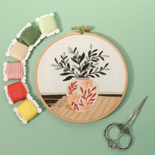 Olive leaf vase embroidery hoop with colorful threads and scissors on green background.