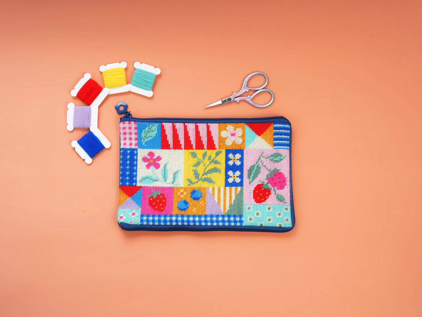 Patchwork Garden Half-Stitch Kit pouch laid flat on a peach background, featuring colorful patchwork floral patterns, embroidery threads, and scissors.
