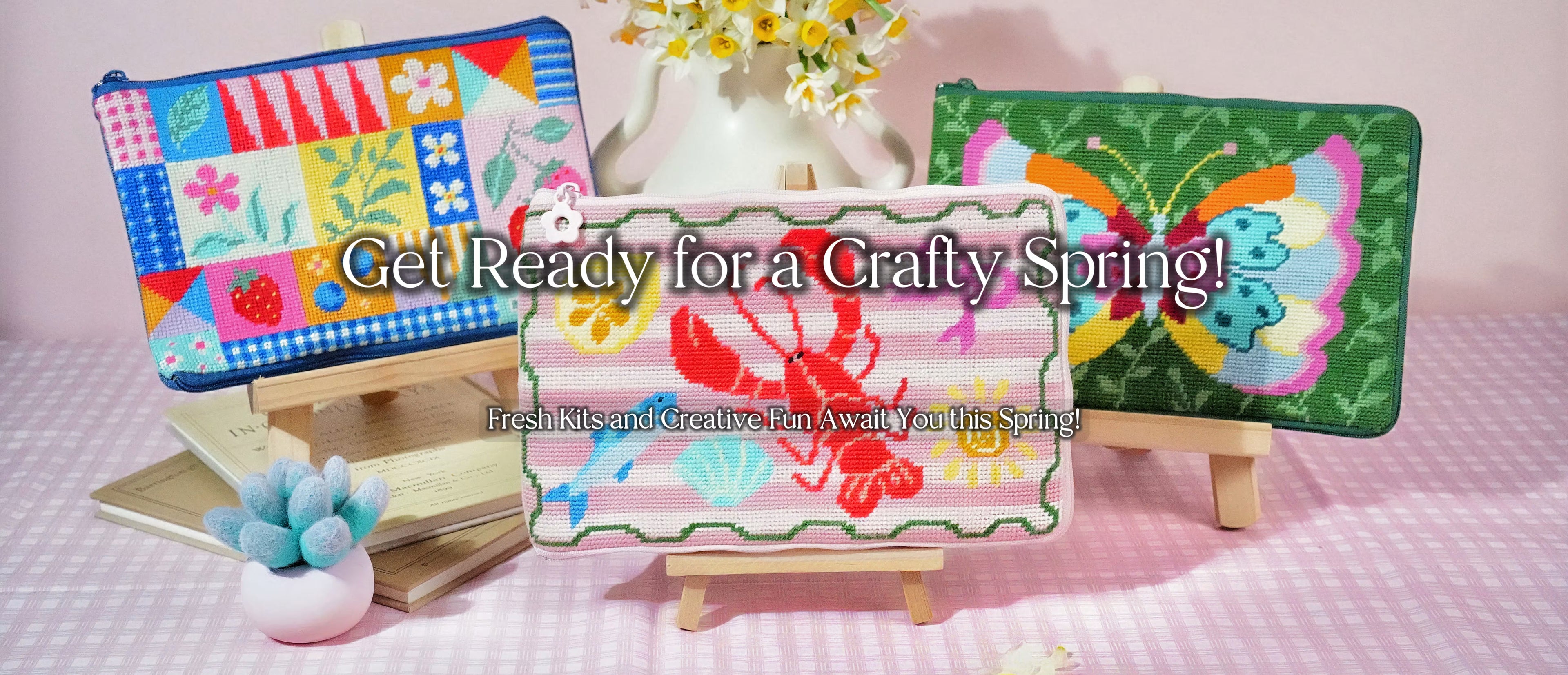 Spring-themed display of Artglee half-stitch kits, featuring colorful stitched pouches on small easels, styled with flowers and soft pastel decor.
