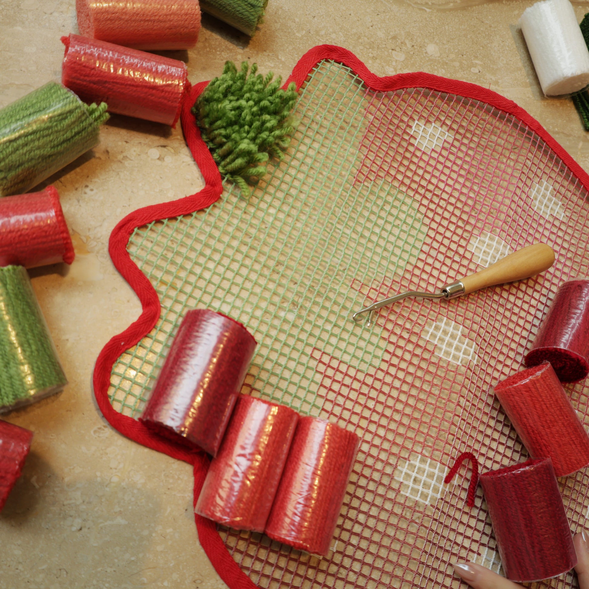 Strawberry-shaped latch hook canvas with green yarn partly filled, surrounded by red and pink yarn rolls and a latch hook tool.