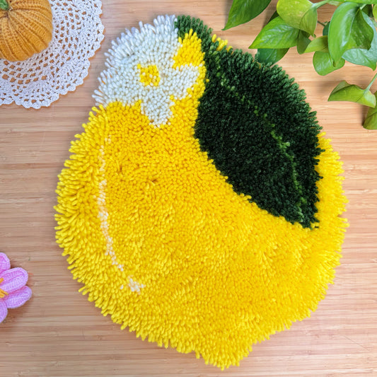Finished latch hook rug shaped like a lemon with green leaf and white flower detail, displayed on a wooden surface with plants and decor.