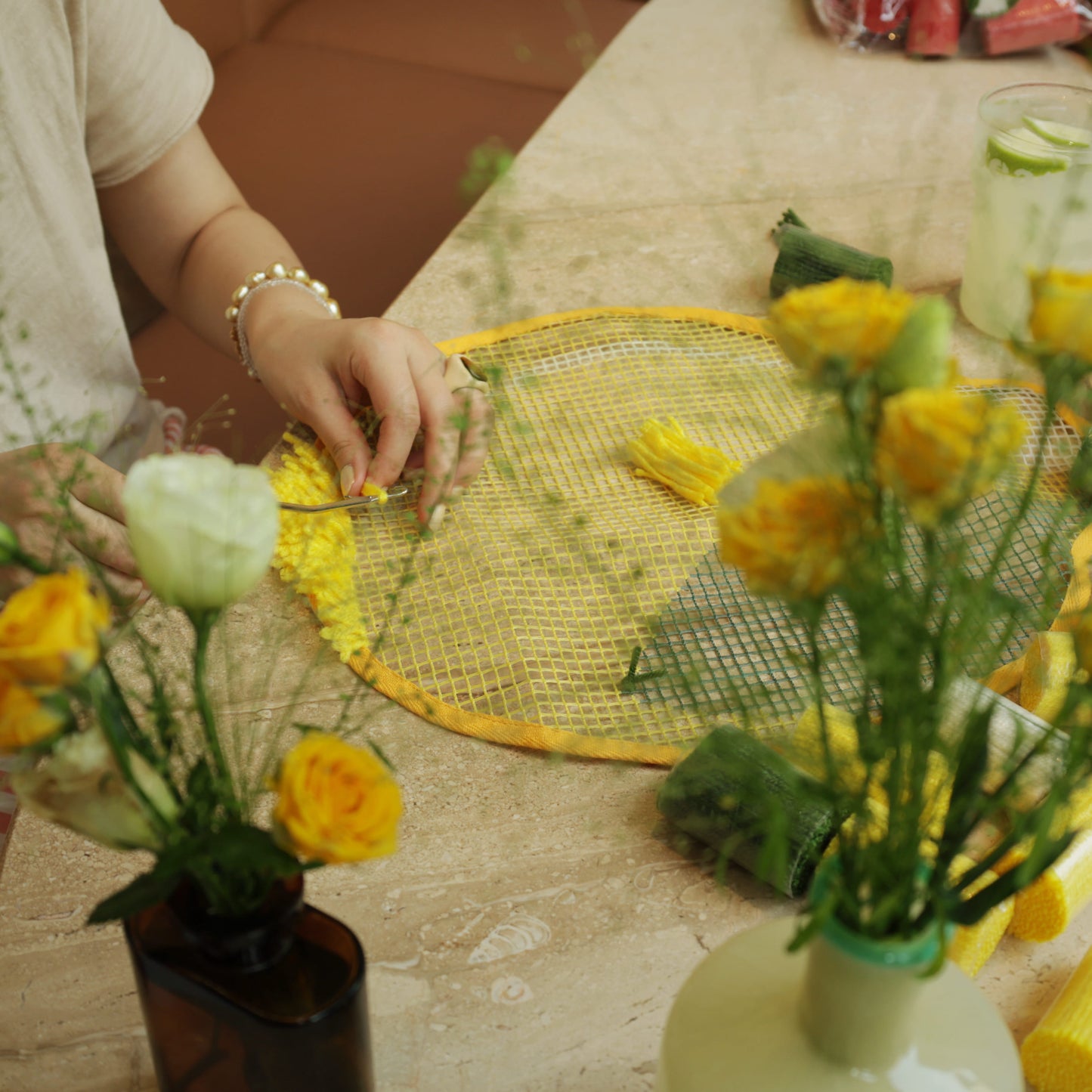 Hands working on the Sunny Lemon latch hook kit, pulling yellow yarn through the mesh canvas at a wooden table.